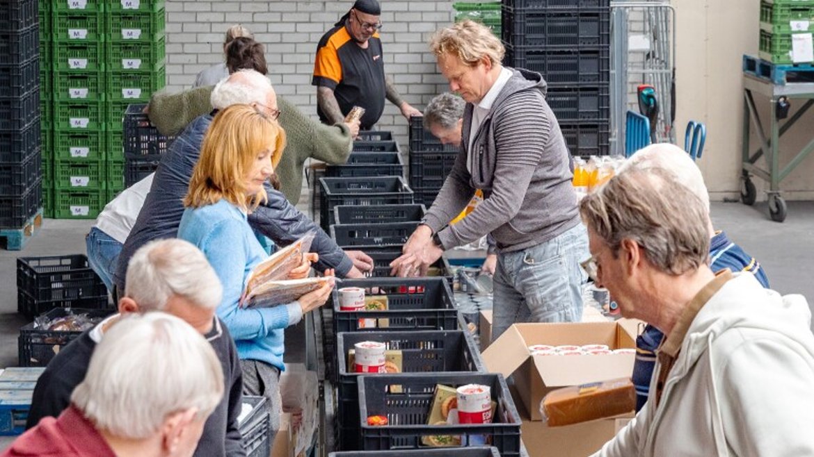 Vrijwilligers sorteren voedsel en goederen in zwarte kratten in een magazijn.